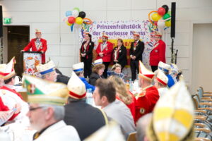 Im Hintergrund Menschen auf einer Bühne, im Vordergrund viele Menschen mit Karnevalskappen
